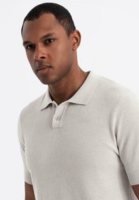 Beige strikket polo skjorte med korte ærmer, der har en klassisk krave og to knapper, glat tekstur og et subtilt mønster.