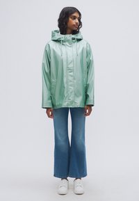 Μέντα πράσινο, γυαλιστερό αδιάβροχο με κουκούλα, μπροστινό κλείσιμο και πλαϊνές τσέπες. Συνδυάζεται με καμπύλα μπλε τζιν και λευκά αθλητικά παπούτσια.