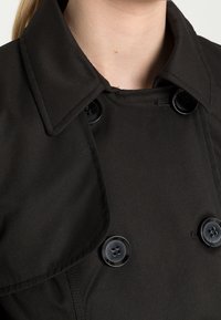 Svart trenchcoat med stora slag, två glansiga knappar och ett slätt, strukturerat tyg. Närbild av krageområdet.