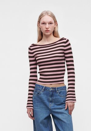 Ung kvinde med langt blondt hår iført en pink og brun stribet langærmet top og løse blå jeans mod en ensfarvet baggrund.