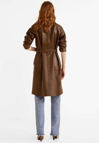 Bruine leren trenchcoat met een ceintuur in de taille, lange mouwen en lengte tot de helft van de kuit. Draag het boven een lichtblauwe spijkerbroek en hakken. Gladde textuur.