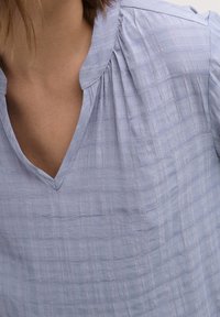 Blouse bleu clair avec col en V, en tissu texturé présentant un motif en grille et de subtiles plis aux épaules.