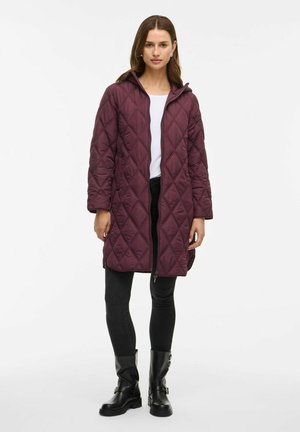 Veste longue matelassée bordeaux avec capuche, dotée d'une fermeture éclair à l'avant et d'échancrures sur les côtés, portée sur un haut blanc et un pantalon noir ajusté.