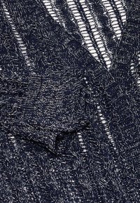 Tengerészeti kötött kardigán, nyitott szövésű, textúrás mintákkal és fémes szálakkal. Laza illeszkedés és leomló fazon jellemzi.