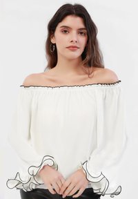Hvit off-shoulder bluse med volangkant i halsen og svart kant, med lange ermer og bølgete kantdetaljer. Glatt tekstur.