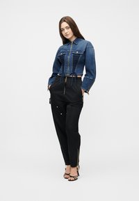 Νεαρή γυναίκα στέκεται με τα χέρια στις τσέπες, φορώντας κοντό μπλε τζιν μπουφάν και μαύρο ψηλόμεσο παντελόνι με φερμουάρ και σανδάλια.