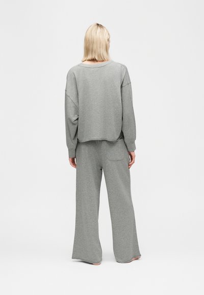 Άτομο με κοντά ξανθά μαλλιά, φοράει έναν φαρδύ γκρι μακρυμάνικο τοπ και παντελόνια με φαρδιά πόδια, στέκεται ξυπόλητο με την πλάτη στο λευκό φόντο.