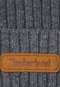Cappello grigio a maglia costellata con un'etichetta in pelle marrone con la scritta "Timberland" in rilievo. Il tessuto strutturato aggiunge profondità al design.