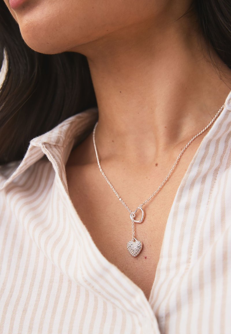 Silver necklace featuring a dangling heart pendant with a diamond-like finish, attached to a chain with a circular link detail.