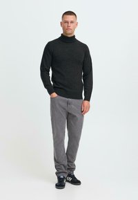 Man staat met één hand in zijn zak, draagt een zwart coltrui, grijze broek en zwarte sneakers met witte strepen.