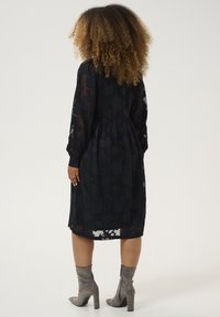Schwarzes transparentes Kleid mit floralem Muster, langen Ärmeln und gerafftem Bund. Kombiniert mit silbernen, verzierten Stiefeletten.