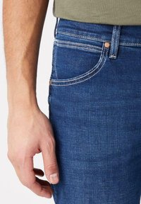 Jeans de mezclilla azul oscuro con costuras claras, que cuentan con cinco bolsillos y un corte clásico. La mano apoyada en el muslo muestra la textura de la tela.