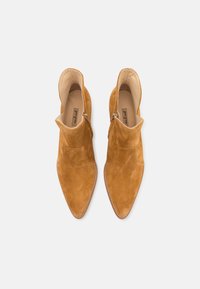 Pár khaki semišových kotníkových bot s špičatými prsty a bočními zipy, zobrazených shora na světlém pozadí.