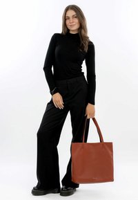Brun læder tote taske med to håndtag, tekstureret overflade, bæres med sort tætsiddende top og vide bukser. Enkel design, ingen synlige mønstre.