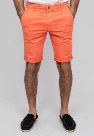Homme portant un short orange vif retroussé, chaussures noires sans lacets, mains dans les poches, debout devant un fond gris uni.