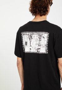 Black cotton t-shirt featuring a white graphic illustration on the back, depicting stylized figures within a bordered frame. Short sleeves.