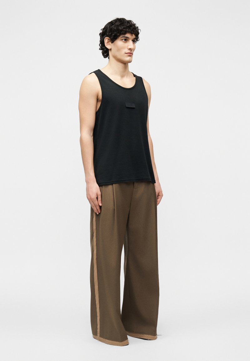 Joven con cabello rizado y oscuro que lleva una camiseta sin mangas negra y pantalones marrones de pierna ancha con rayas laterales beige, de pie contra un fondo liso.