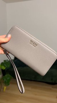 Γκρι πορτοφόλι από συνθετικό δέρμα με υφή, κλείσιμο με φερμουάρ, λουράκι για τον καρπό και χρυσή ανάγλυφη στάμπα "GUESS" στο μπροστινό μέρος.