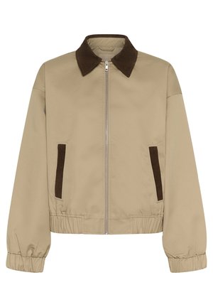 Frau trägt beige Jacke, weißes Top, langen dunklen Jeansrock und flache Schuhe mit Zebra-Streifen und schwarzen Riemen, steht mit einer Hand in der Tasche.