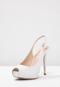 Scarpa con tacco alto bianca, slingback, con punta aperta, esterno in pelle lucida e dettaglio in metallo sul tacco. Presenta una sottile cintura regolabile con fibbia.