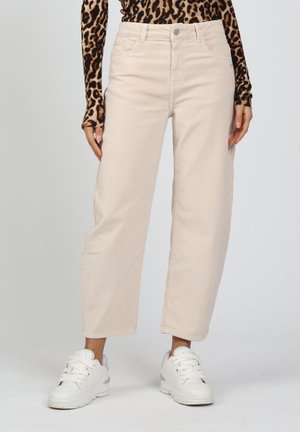 Person trägt beige Cordhosen mit weitem Bein, weiße Turnschuhe und ein langärmliges Oberteil mit Leopardenmuster, steht vor einem schlichten Hintergrund.