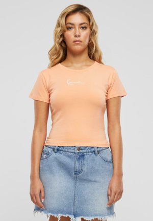 T-shirt ajusté à manches courtes de couleur pêche avec col rond et logo blanc sur la poitrine, associé à une jupe en denim délavé avec un ourlet effiloché.