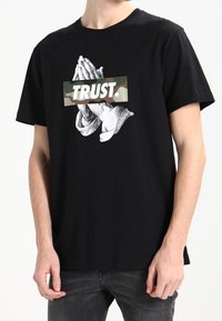 Sort bomulds T-shirt med et grafik af bedende hænder på en camo-baggrund og ordet "TRUST." i fede hvide bogstaver.