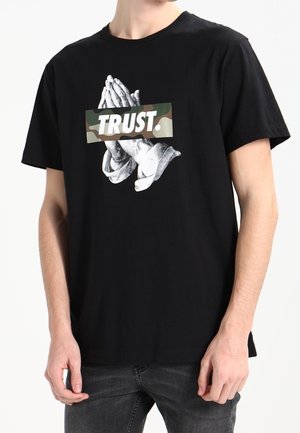 Camiseta de algodón negra con un gráfico de manos en oración sobre un fondo de camuflaje y la palabra "TRUST." en letras blancas y en negrita.