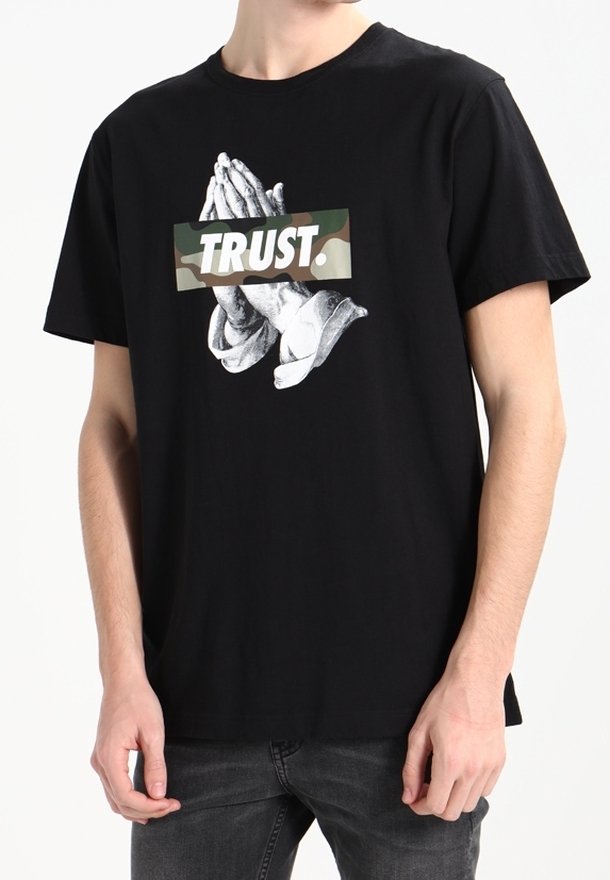 Sort bomulds T-shirt med et grafik af bedende hænder på en camo-baggrund og ordet "TRUST." i fede hvide bogstaver.