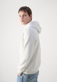 Sudadera blanca de material suave con un bolsillo frontal, puños y dobladillo de canalé; combinada con jeans azules desgastados. Diseño simple y casual.