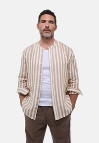 Chemise à rayures beige et blanche en lin avec un col montant, manches longues retroussées, portée sur un T-shirt blanc.