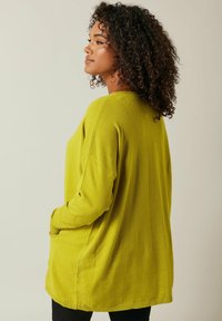 Langärmliger Pullover in lässiger Passform aus leuchtend gelb-grünem Strick, mit seitlichen Taschen und geripptem Saum, seitlich gegen einen neutralen Hintergrund gezeigt.