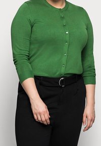 Groen vest gemaakt van een zachte, gestructureerde stof met een ronde halslijn en knoopsluiting. Gecombineerd met zwart, hooggetailleerd broek en een riem.