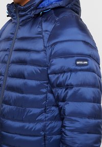 Donkerblauwe gewatteerde pufferjas met een doorgestikte textuur. Heeft een hoge kraag, ritssluiting en een klein logoplaatje op de mouw. Het materiaal lijkt glanzend.