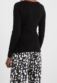 Schwarzes langärmliges Oberteil mit strukturiertem Rippdesign und weitem Ausschnitt, kombiniert mit schwarz-weißen gemusterten Hosen mit abstrakten Formen.
