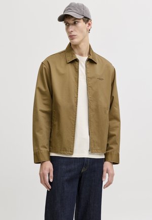 Mlad moški, ki nosi rjavo jakno z zadrgo, belo srajco, temne kavbojke in sivo baseball kapo, gleda levo na enostavni podlagi.