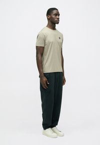 Bézs színű pamut póló kis logóval a mellkason, sötétzöld cargo nadrággal és fehér tornacipővel párosítva. A modell oldalnézetben áll.