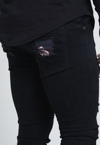 Black denim jeans with a slim fit, featuring a distressed back pocket and a floral patch detail. The fabric has a slightly textured appearance.