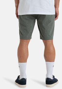 Pantaloni corti di tessuto verde scuro con un motivo testurizzato, lunghi fino al ginocchio, abbinati a calze bianche a coste e scarpe da ginnastica blu navy, visti da dietro.