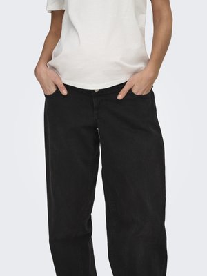 Personne portant un pantalon noir ample et un t-shirt blanc, les mains dans les poches avant, sur un fond uni.