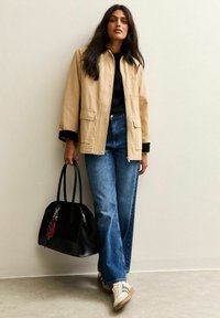 Beige canvas jas met knoopsluiting en voorzakken, gecombineerd met blauwe uitlopende jeans. Zwarte handtas met franjes en witte sneakers.