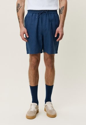 Man draagt marineblauwe short met trekkoord, witte sneakers en marineblauwe sokken, staat met ontspannen armen langs het lichaam.