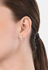 Orecchini d'argento con un design a goccia, caratterizzati da una finitura liscia e un piccolo gruppo di strass trasparenti. Ambientati su uno sfondo neutro.