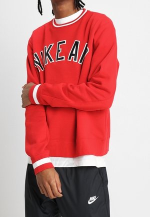 Rød sweatshirt med sort "NIKE" bogstavtryk. Har hvid ribbet krave og manchetter. Bæres over en hvid skjorte, parret med sorte bukser.