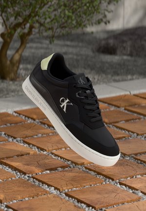 Black Calvin Klein sneaker with white sole and logo on side, floating above wooden square tiles with small gray pebbles in outdoor setting.
