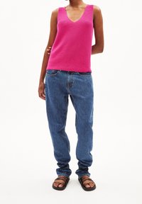 Kvinna iklädd en ljusrosa ärmlös stickad topp, lösa blå jeans och svarta sandaletter med remmar, stående mot en vit bakgrund.