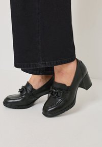 Črne usnjene čevlje s peto, ki vsebujejo naglas verige, okroglo konico in teksturirano podplat. Imajo gladko površino in rahlo dvignjeno peto.