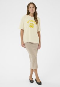 Femme portant un T-shirt crème surdimensionné avec le texte jaune "ST TROPEZ" et un graphique de cerises, une jupe midi beige et des ballerines noires.