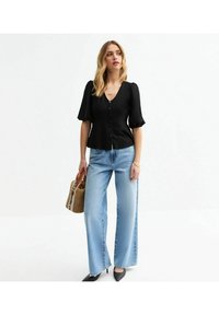 Svart knappad topp med korta puffärmar, ihop med ljusblå vida jeans och svarta spetsiga klackar, som håller en vävd handväska.
