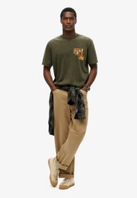 Oliwkowa bawełniana koszulka z grafiką na piersi, w połączeniu z beżowymi spodniami sztruksowymi i czarno-zieloną koszulą w kratę zawiązaną w talii.